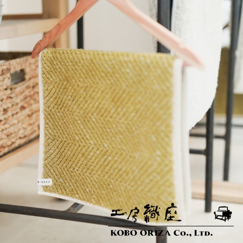 一塊黃色棉麻混紡人字紋快乾浴墊，掛在木質衣架上，背景有編織籃和白色毛巾。