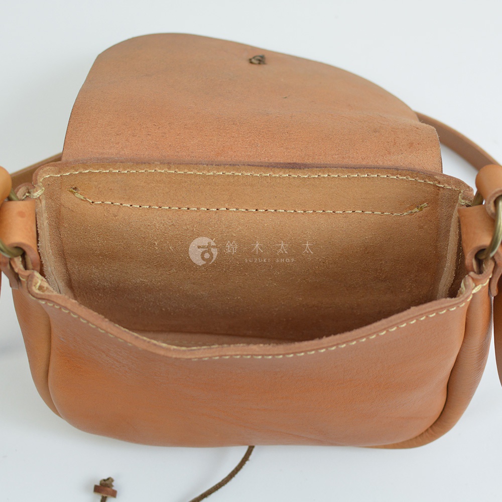 職人手作的淺棕色皮革圓形翻蓋肩背包，內部空間寬敞，側邊有金屬扣環，背帶為細長皮革繩。