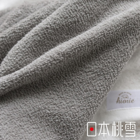 一條觸感柔軟的灰色今治浴巾，材質為純棉，表面有細緻的毛圈紋理，部分區域有摺疊效果，右下角可見品牌標籤「hiorie」及「MADE IN JAPAN」字樣，以及中文「日本桃雪」。
