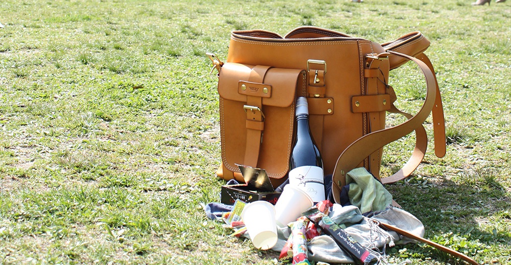 職人手作皮革大型三用旅行後背包，淺棕色，配有金屬扣環和拉鍊，包包旁有酒瓶、紙杯和零食。