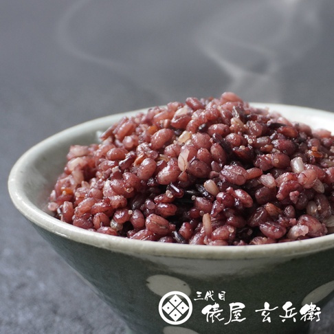 一碗熱氣騰騰、呈現深紫紅色的日本國產十六穀米，盛裝在帶有圓點花紋的綠色陶碗中，米飯顆粒飽滿且質地豐富，碗身右下角印有三代目俵屋玄兵衛的品牌標誌與書法字樣，整體散發出健康且美味的視覺感受。