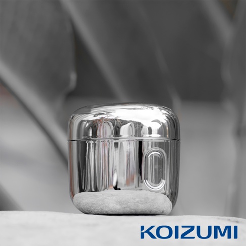 一顆置於石塊上的Koizumi日本品牌超迷你刮鬍刀，擁有具備鏡面金屬光澤的圓潤機身，表面清晰反射出周圍環境，下方邊緣處設有簡約的橢圓形凹槽細節。