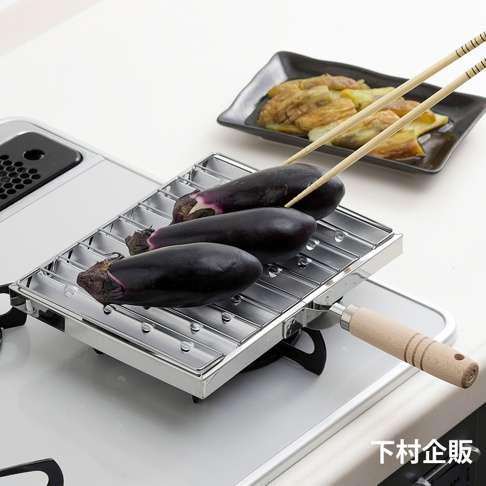 不鏽鋼材質的瓦斯爐專用炭烤風萬用燒烤盤，架上擺放著三根紫色的茄子，一雙木製筷子夾著其中一根茄子，準備進行燒烤。