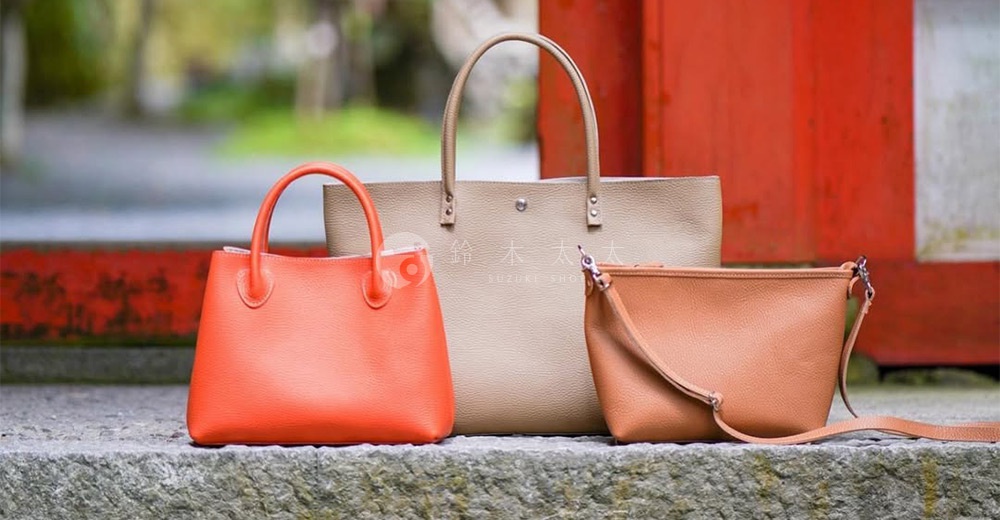 三款皮革手提托特包，分別為亮橘色、米白色和焦糖色。亮橘色包為手提款，米白色包為大型托特款，焦糖色包為斜背包款，均展示皮革紋理。