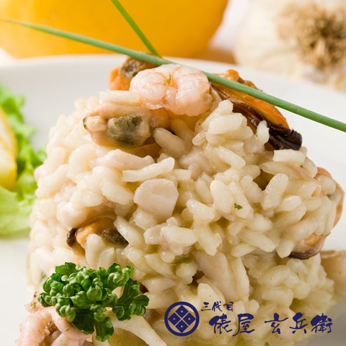 一盤飽滿的燉飯，米飯呈現米白色，混合著鮮蝦、淡菜和切塊的魚肉，上方點綴著青蔥和綠葉蔬菜，背景有模糊的黃色物體和蒜頭。