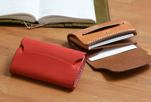 兩個職人手作皮革迷你卡片收納夾，一個為鮮紅色，另一個為淺棕色。紅色的收納夾閉合，展示其光滑的皮革表面。棕色收納夾打開，露出內部夾層，可容納卡片。