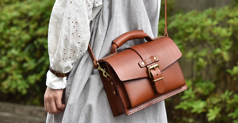 一個棕色皮革迷你單肩包，配有頂部提把、可調節肩帶、黃銅色扣環和品牌標誌。包包採用縫線工藝製作。