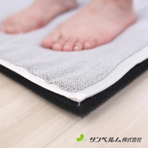 一雙赤腳踩在淺灰色毛圈材質的浴墊上，該浴墊底部採用黑色的透氣防滑網狀結構設計，並放置在木質地板上，角落處印有淺綠色圖標與サンベルム株式会社的品牌名稱。