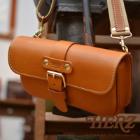 職人手作皮革單扣經典斜背包，採用鞣製皮革製成，顏色為溫暖的棕褐色，配有金色金屬扣環和可調節的帆布與皮革拼接背帶。