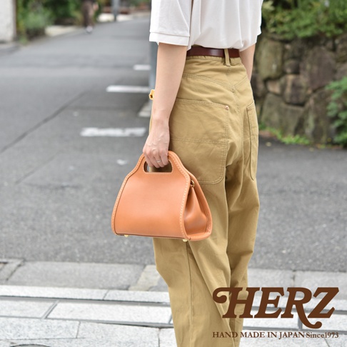 職人手作皮革單肩兩用拱型手袋，採用光滑皮革製成，呈現自然的棕褐色調。手袋設計簡潔，帶有雙提把和可調節的單肩背帶。精緻的縫線和金屬配件增添質感。