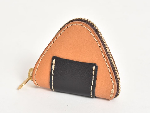 手工皮革零錢包，呈三角飯糰造型，主體為淺棕色皮革，正面中間有一塊黑色皮革裝飾，並以白色縫線點綴，搭配金色拉鍊和鑰匙圈。