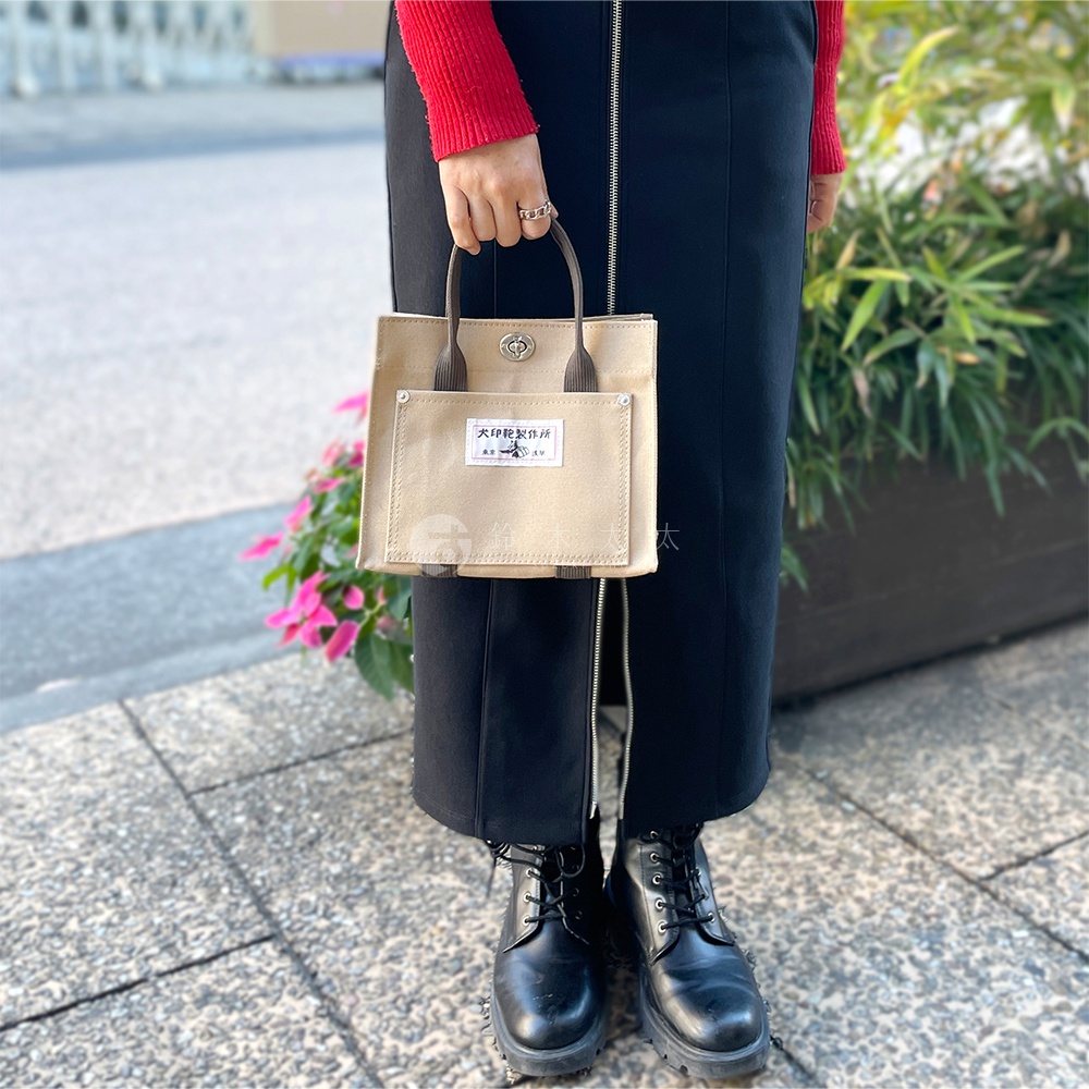 米色純棉帆布迷你托特包，有深棕色提把和外層口袋，上面印有標籤，由手持。