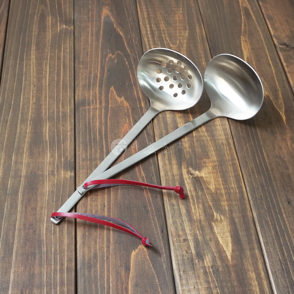 兩支不鏽鋼湯勺，一支為帶有圓孔的濾網勺，另一支為實心湯勺，均配有紅色皮革繫繩。