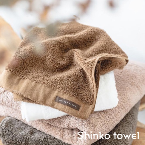 MAISON BLANC 輕奢柔膚匹馬棉毛巾，堆疊展示，包含棕色、白色、淺粉色和灰色。頂部毛巾為棕色，質地柔軟蓬鬆，邊緣有織帶標籤。