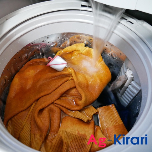 洗衣槽內，一件橘黃色毛衣正在水中浸泡，白色網狀的洗衣槽去污劑綁在毛衣上，水流從上方注入。