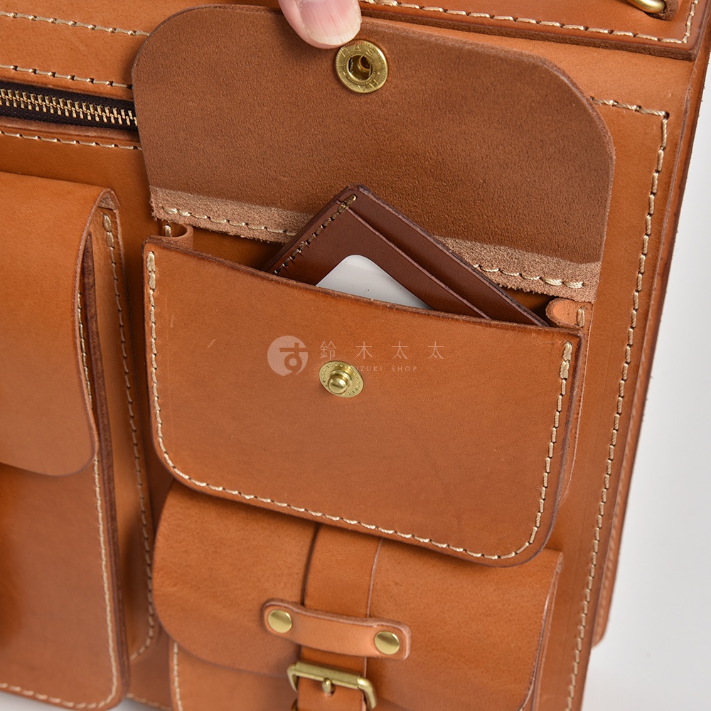 職人手作皮革後背包，呈現自然棕色，以精緻縫線勾勒出多個口袋結構，包含一個帶有銅釦的翻蓋小袋，內部可見卡片等小物。