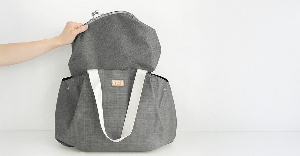 手拿著灰色的 AYANOKOJI Sarei 機能布兩用口金托特包，包包上有白色織帶和品牌標籤