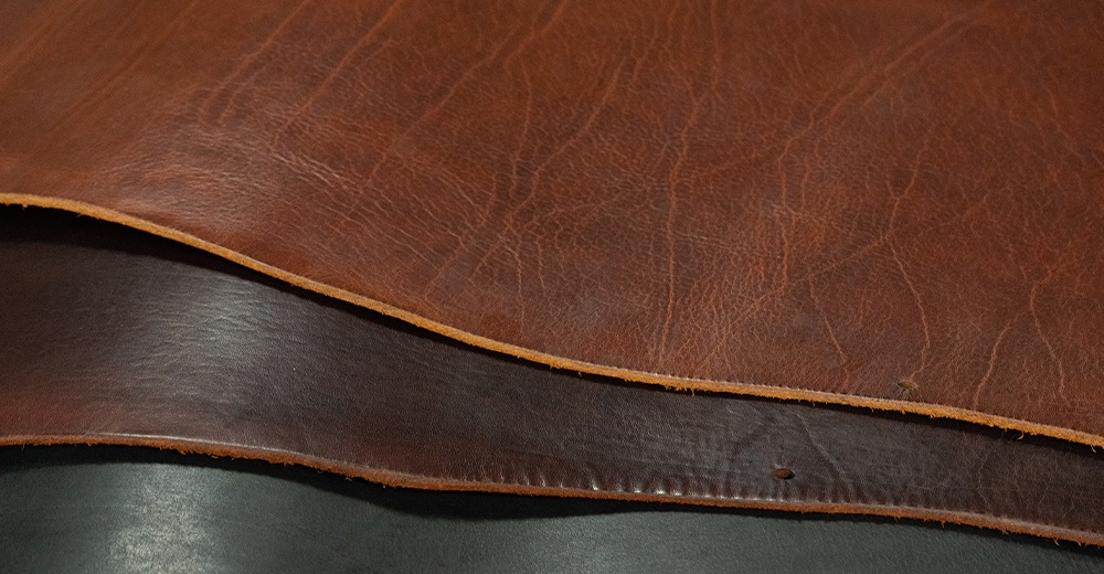 一張咖啡色皮革的特寫，其中有兩塊皮革交疊，上面的一塊顏色較深，呈現自然的紋理，下面的一塊則呈現較深的棕色。