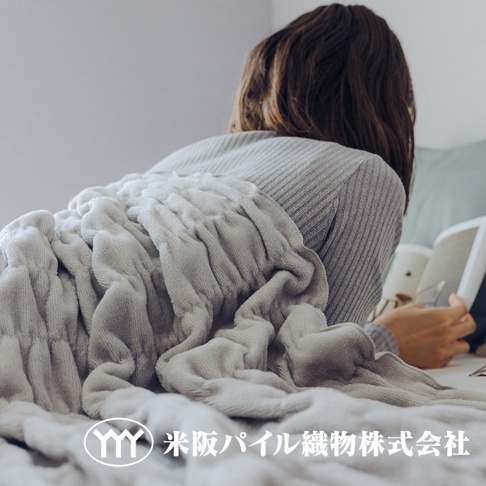 一件淺灰色、蓬鬆柔軟的毛毯，帶有褶皺設計，覆蓋在一位女性身上，她正躺著看書。