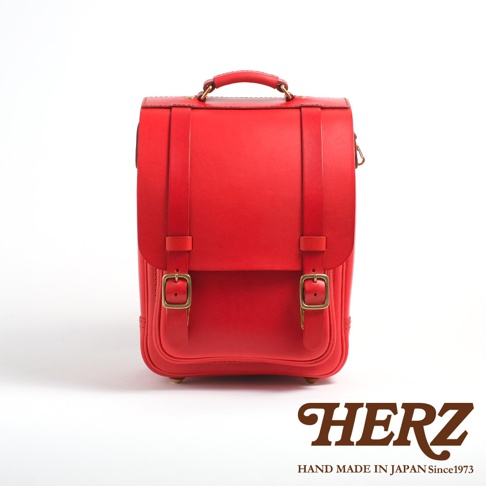 紅色皮革小學生書包，配有兩個黃銅色插扣和提把，手工製作於日本。