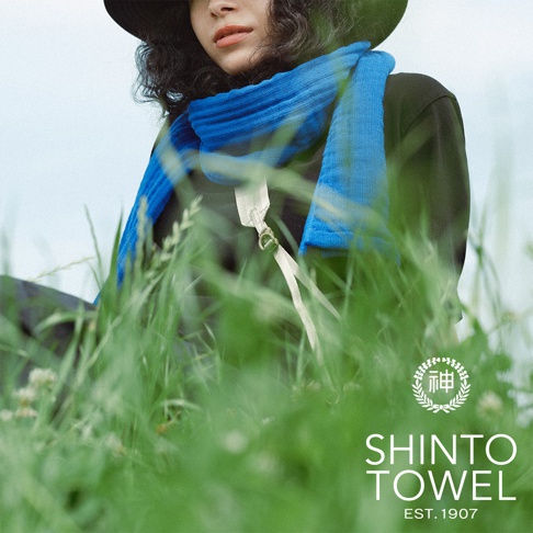 一名女子戴著黑帽，身穿深色上衣，圍著一條鮮藍色的針織圍巾，圍巾上有白色掛繩和金屬扣環。背景為綠色草地與淡藍色天空。