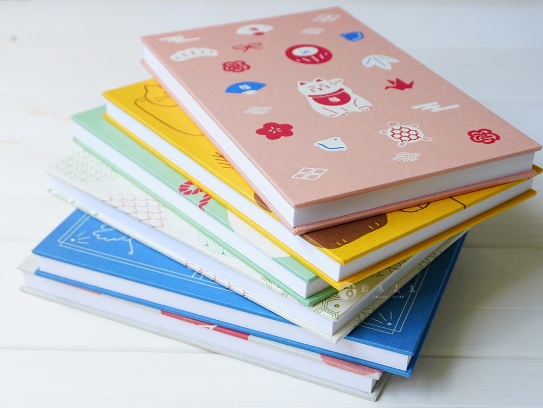 堆疊的御朱印帳，最上方粉色封面印有招福貓、花朵、烏龜等日式圖案。下方依序為黃色、綠色、米白色、藍色等不同封面設計。