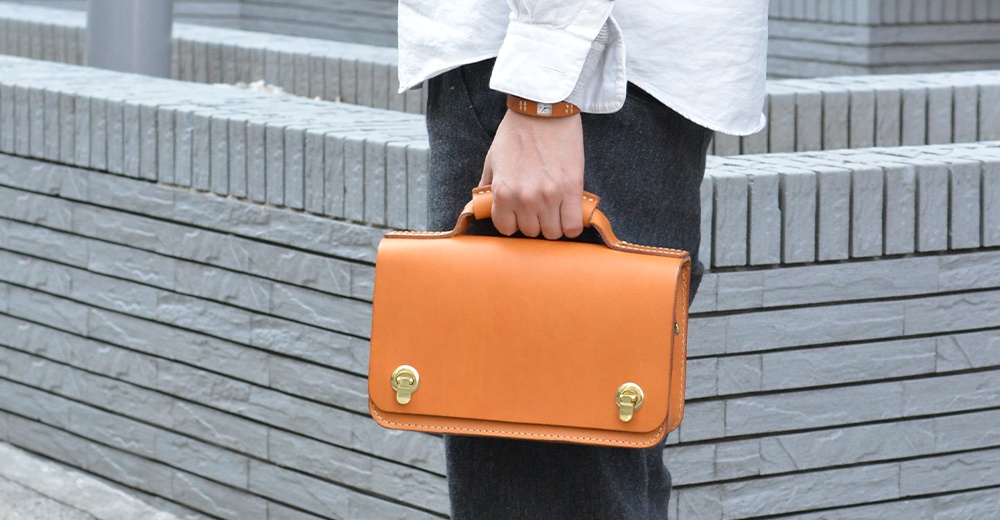 職人手作的經典兩用雙扣迷你單肩包，採用橘棕色皮革製成，配有金色旋轉鎖扣和提把。