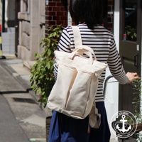 米白色帆布後背包，雙肩帶可調節，正面有兩個立體口袋，上方有提把，搭配藍色長裙與條紋上衣，背景為巷弄街景。