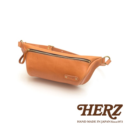職人手作皮革輕便斜背包，材質為天然皮革，顏色為淺棕色。包包設計簡潔，有一個拉鍊主袋，配有可調節的皮質背帶和金屬扣環。包包正面有一個壓印的品牌標誌。
