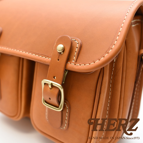 日本HERZ職人手作駝色皮革雙扣郵差包，特寫展示包包的縫線細節、金屬釦環與皮質的質感。