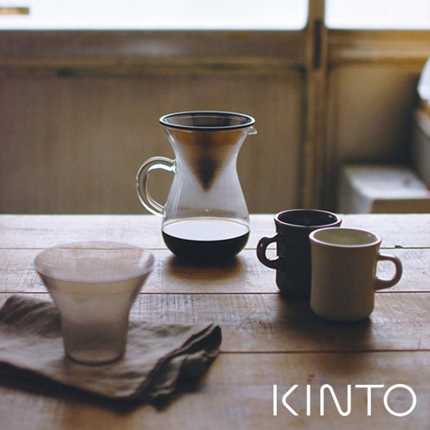 玻璃咖啡分享壺，配有濾杯和兩個馬克杯，分別為深灰色和米白色，置於木質桌面上的麻布上。