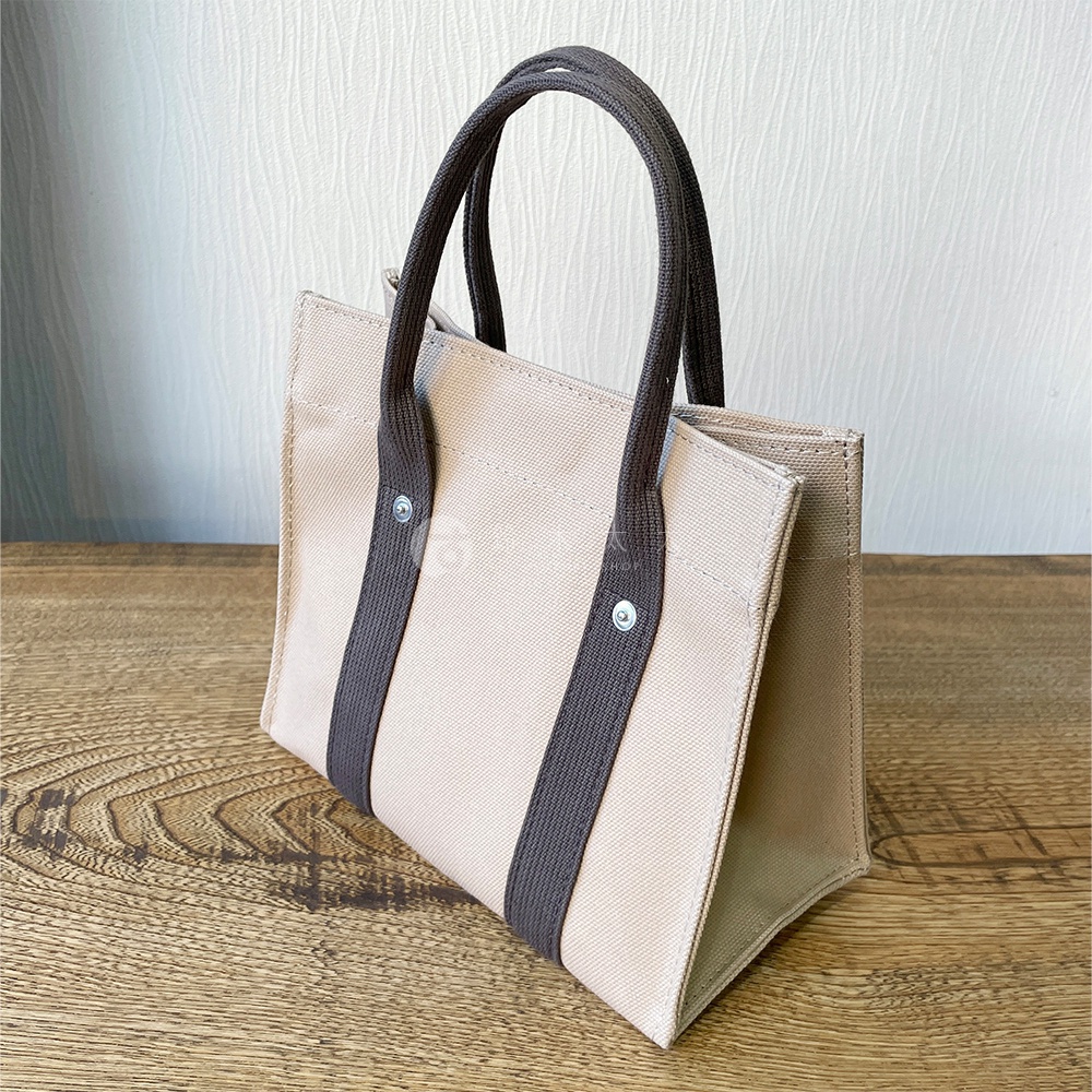 米白色純棉帆布迷你托特包，配有深棕色織帶提把，包身兩側有織帶裝飾，以金屬釦固定，風格簡約。
