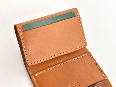 職人手作皮革三折式迷你短夾，採用淺棕色皮革，內裡有卡槽，卡夾內插著一張綠色卡片，車縫線為米白色。