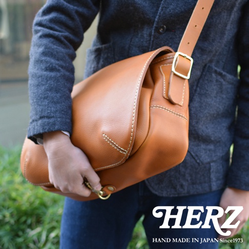 棕色皮革復古翻蓋郵差包，包身線條簡潔，以白色縫線裝飾，配有可調節的皮革肩帶及金色金屬扣環，由人員側身展示。