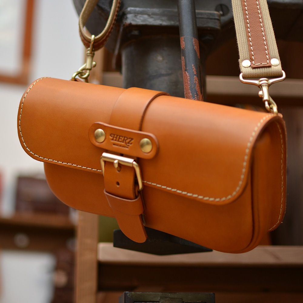 淺棕色皮革單扣經典斜背包，配有可調節帆布和皮革肩帶，使用白色縫線裝飾，風格簡約。