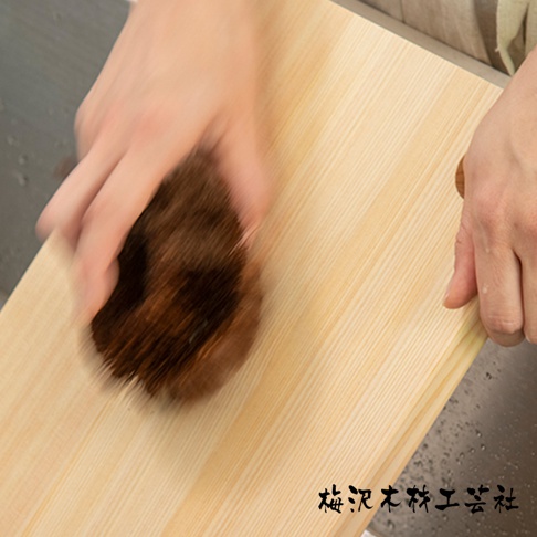 一隻手正拿著圓形棕色天然棕櫚毛刷，在淺木色木板上刷動。