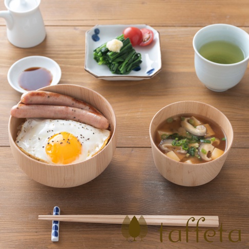 楓木碗裝有白飯、煎蛋和兩根香腸。右側的楓木碗裝有味噌湯，內有豆腐、香菇和蔥花。背景有醬油碟、裝有菠菜、番茄和蘿蔔泥的小盤，以及一杯綠茶。