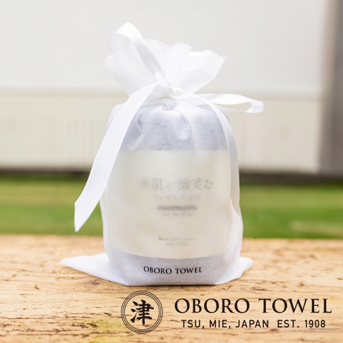 白色半透明網狀束口袋包裝著一條淺灰色毛巾，上面印有日文「潤澤素肌微笑速吸毛巾」及英文「OBORO TOWEL」。束口袋以白色緞帶繫緊，整體呈圓柱狀，放置於木質桌面上，背景為模糊的綠色草地與白色牆壁。