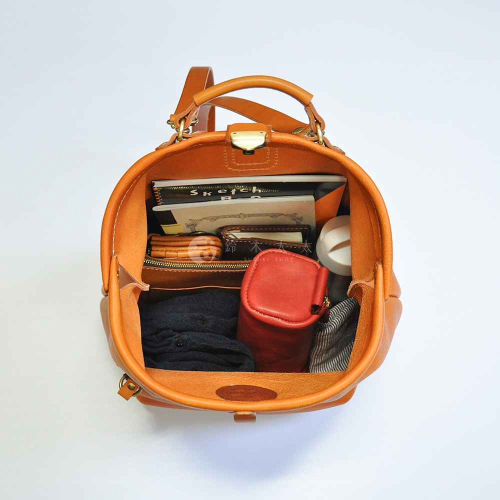 職人手作皮革輕旅行後背包 R-144，展示橘棕色皮革材質，內部裝有筆記本、錢包、紅色小收納包、黑色衣物及條紋布料物品。