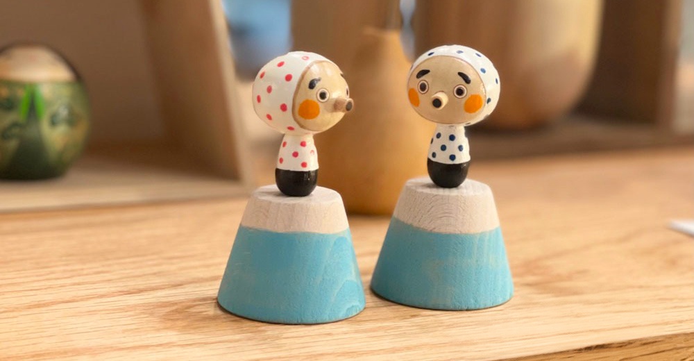 兩尊木芥子開運擺飾，一尊頭戴紅點白頭巾，另一尊頭戴藍點白頭巾，均有圓形臉龐、突出鼻頭和圓點裝飾的身體，基座為木質，上半部為淺木色，下半部為淺藍色。