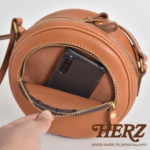 職人手作皮革小圓包，展現溫潤的焦糖棕皮革質感，外露的車縫線與圓潤的包身設計，開口處拉鍊內可見一支手機，展現其收納功能。
