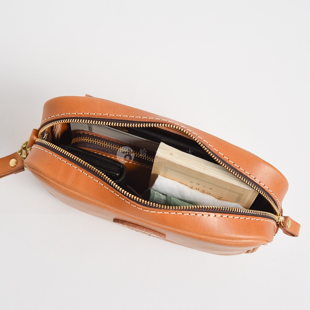 棕褐色皮革手提包，拉鍊部分打開，內有錢包、書本和綠色織物。包包採用縫線裝飾，配有可拆卸的扣環。