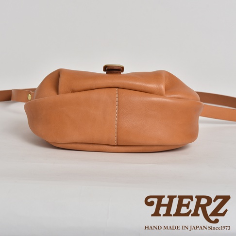 HERZ 品牌標誌與 MADE IN JAPAN Since1973 字樣，背景為一只手工製作的棕褐色皮革斜背包的底部特寫，呈現皮革的紋理與縫線細節。