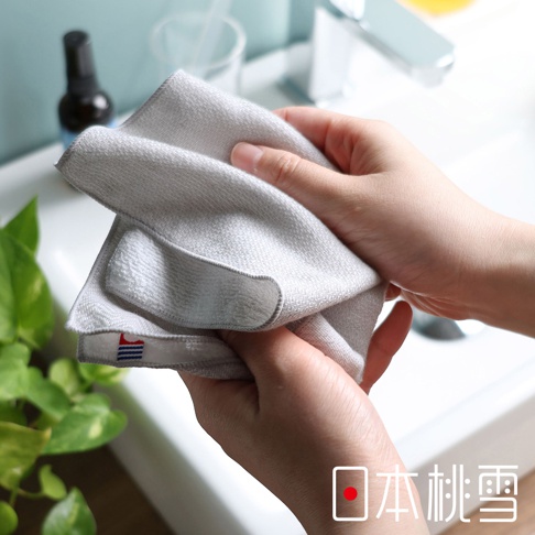 一雙手正拿著一條淺灰色的人字紋紗布方巾，毛巾一角印有藍色和紅色的條紋標籤，下方有「日本桃雪」的日文標誌。