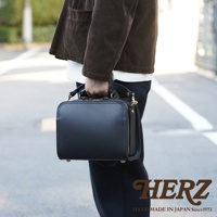 職人手作黑色皮革兩用方形單肩包，包身結構方正，配有金色拉鍊與釦環，展現簡約俐落的設計感。