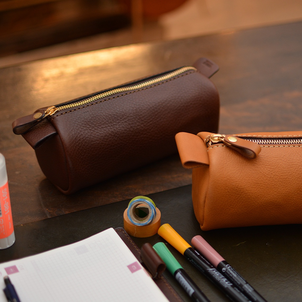 兩款職人手作皮革多用途收納筆袋，分別為深棕色和淺棕色，皆採用粒面皮革材質，配有金色拉鍊。筆袋造型簡約，細節處可見皮革提把和縫線。