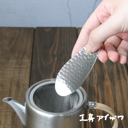 一只手握著錘紋銀色不鏽鋼茶勺，勺面有蜂窩狀紋理，勺尖朝向一個銀色帶有細網濾網的茶壺。