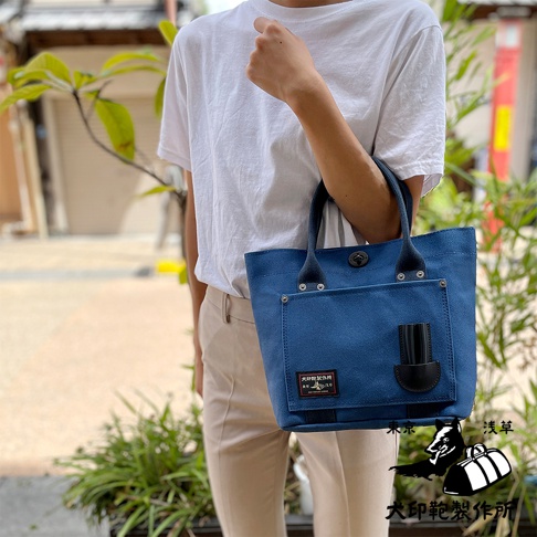 藍色帆布水桶托特包，有黑色皮革提把與細節，包包正面有方形外袋，並附有品牌標籤。