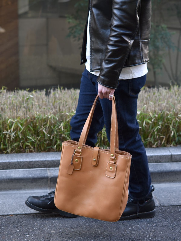 淺棕色皮革托特包，配有兩個頂部提把、金屬原子釦和皮革飾帶。