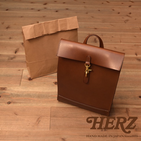 棕色皮革製成的紙袋風後背包，具有翻蓋式設計和黃銅扣環，旁邊有一個類似的棕色紙袋。